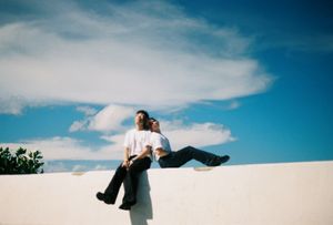 Two people sitting together on a white wall under a bright blue sky with fluffy clouds on a sunny day.