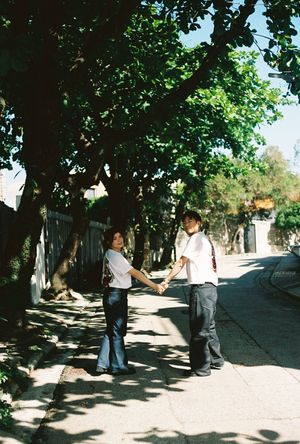 A couple holds hands on a sunny tree-lined sidewalk, both wearing white shirts and dark pants.