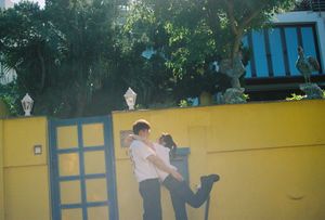 Two people embracing near a bright yellow wall with a blue door, surrounded by lush green trees.