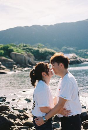 A young couple in matching white t-shirts share a romantic moment by a rocky shoreline with mountains in the background.