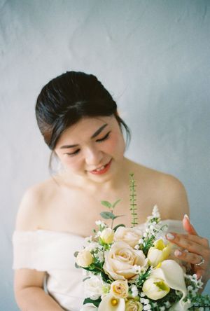 Bride in off-shoulder white gown smiling softly while holding elegant bouquet of cream roses and greenery.