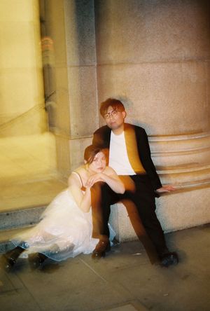 Couple in wedding attire sitting against a stone column at night, woman in white dress leaning on man in black suit.
