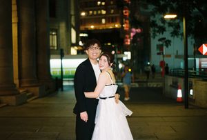 Couple in formal attire embracing on a city street at night, woman in white dress and man in black suit.
