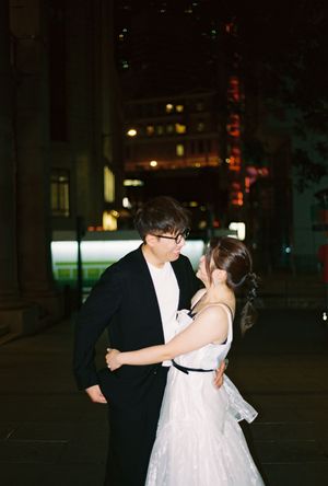 Couple in wedding attire sharing a romantic moment on a city street at night, with glowing buildings in the background.