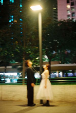 Couple in formal attire gazing up at a street lamp at night with city buildings in the background.