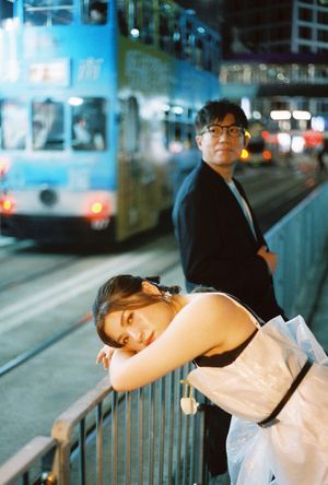 Couple posing at night near a blue tram on a city street, woman leaning on railing in white dress.