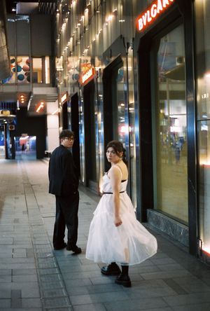 Couple in formal wear on urban sidewalk at night near Bvlgari store, woman in white dress, man in black suit.