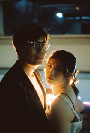 A couple poses dramatically in dim light, the man wearing glasses and a dark jacket, the woman leaning close behind him.
