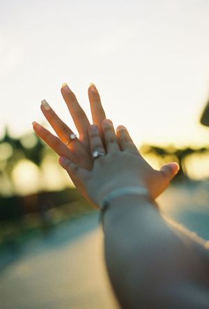 Close-up of a hand wearing a diamond engagement ring and bracelet raised against a warm golden sunset background.