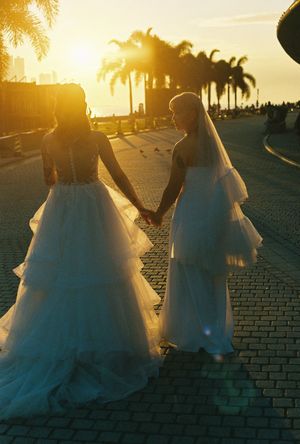 Two brides in white gowns hold hands at sunset near palm trees on a cobblestone plaza.
