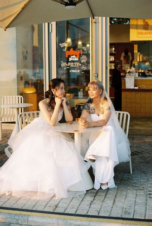Two brides in white wedding dresses sitting together at an outdoor cafe table near a gelato shop.