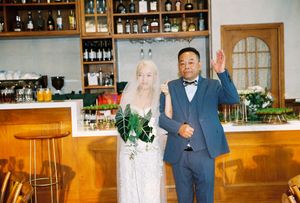 Bride and groom posing together in front of a bar, bride holding tropical greenery bouquet, groom in blue suit waving.