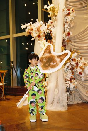 Young child in green printed jumpsuit holding a large cat face cutout sign at a floral decorated event venue.