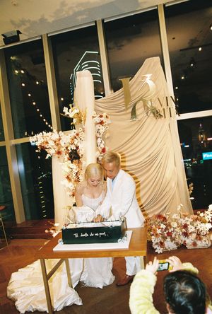 Bride and groom cutting wedding cake at elegant reception with floral arrangements and city skyline backdrop at night.