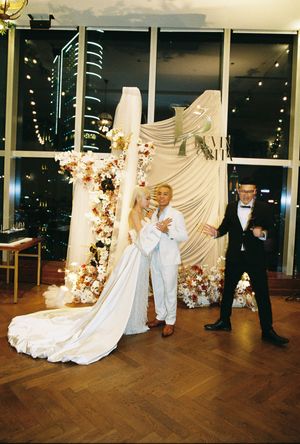 Bride in white gown with groom in white suit posing at floral wedding backdrop, guest in tuxedo nearby.