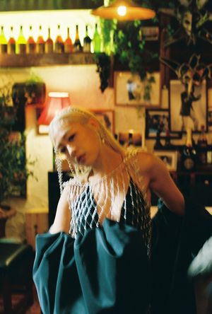 A blonde woman in a teal dress with beaded net accessories poses in a warmly lit, eclectic bar setting.