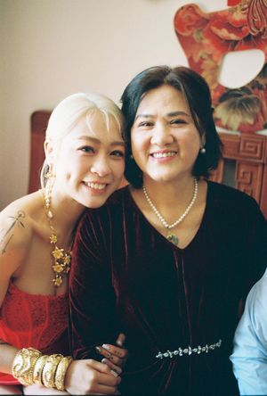 Two smiling women pose together, one wearing gold jewelry and red dress, other in black velvet top with pearl necklace.