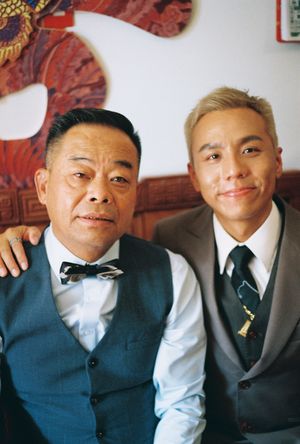 Two men in vests and ties pose together, one wearing a bow tie, smiling at the camera in a formal setting.
