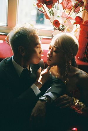 A couple shares an intimate moment surrounded by red roses near a window, the woman adorned in gold jewelry.