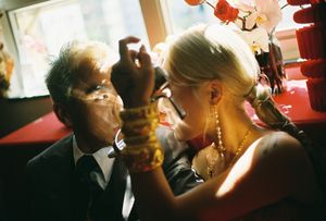 An older man and a blonde woman share an intimate moment at a restaurant, adorned with gold jewelry and elegant attire.
