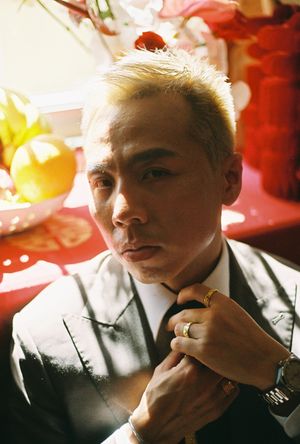 Man with blonde hair adjusting tie, wearing suit and gold ring, with red festive decor in background.
