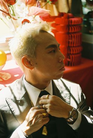 Man with bleached hair adjusting tie, wearing suit and gold rings, with red decorations in background.