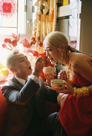 A couple in traditional red wedding attire shares tea during a Chinese wedding ceremony, smiling joyfully.