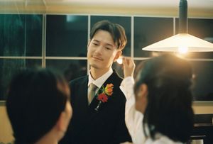 Groom in black vest with floral boutonniere adjusting earring indoors under warm lamp light.