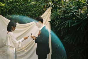 Bride and groom exchanging vows outdoors with white draped backdrop and lush green foliage.