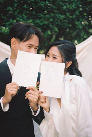 Couple holding hand-drawn portrait sketches at outdoor wedding ceremony with white drape backdrop.