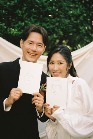 Smiling couple holding hand-drawn portrait sketches at an outdoor wedding ceremony, man in black suit, woman in white dress.