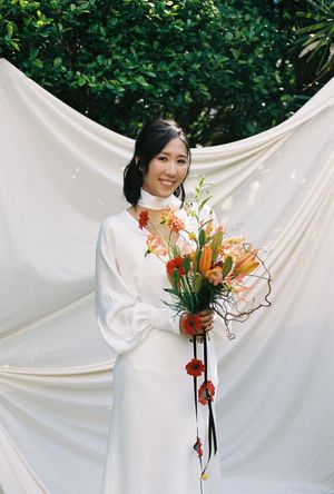 A bride in a white dress holds a vibrant orange floral bouquet, smiling before a white draped backdrop.