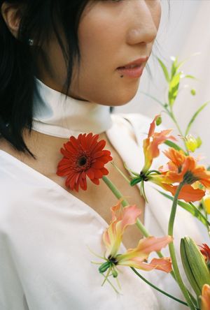 A young Asian woman in white holding vibrant red gerbera daisy and orange lily flowers close to her chest.