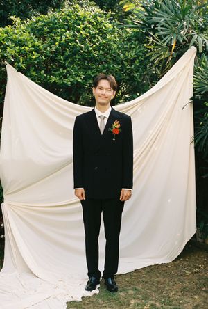 A groom in a black suit with floral boutonniere stands smiling before a white draped backdrop outdoors.