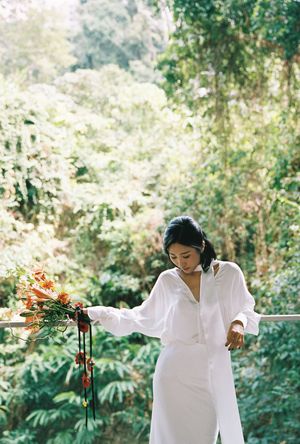 Woman in white dress holding orange flowers on a balcony surrounded by lush green tropical forest.
