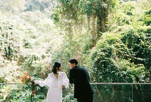 A bride in a white dress and groom in black suit stand on a balcony surrounded by lush green tropical forest.
