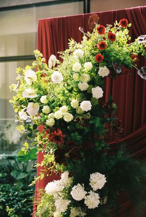 Lush floral arrangement with white carnations, red gerberas, and green foliage against a red draped backdrop.