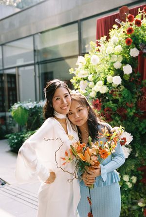 Two smiling women pose together holding orange floral bouquet in front of white and red flower arrangement.