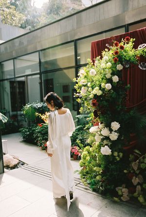 Woman in white dress standing beside a lush floral arch with red and white blooms in a glass courtyard.