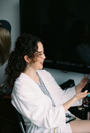 A smiling woman with curly dark hair in a white robe sits at a vanity, gesturing with her hands during bridal preparations.