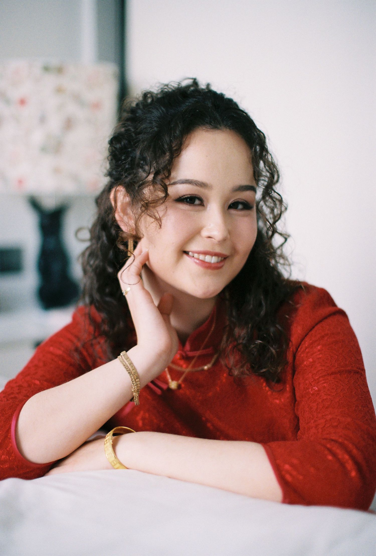 Young woman in red dress with curly hair and gold jewelry smiling warmly while posing indoors.