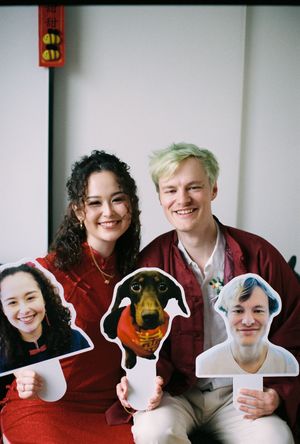 Smiling couple in red outfits holding cutout photos of faces and a dog at a festive gathering.