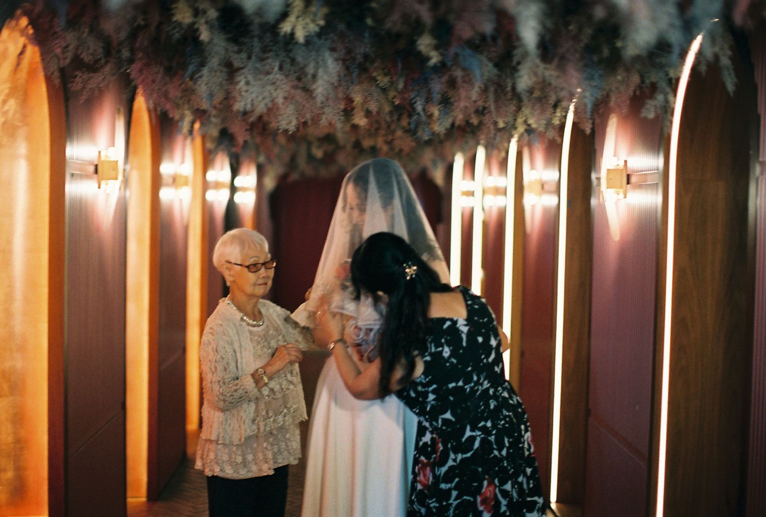 Bride in white gown and veil being assisted by two women in a warmly lit venue decorated with floral arrangements.