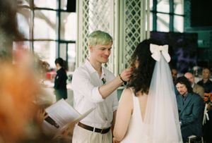 Groom with green hair adjusts brides veil during outdoor wedding ceremony with guests.
