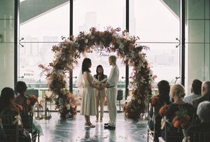Couple exchanging vows under a floral arch at an indoor wedding ceremony with guests seated nearby.