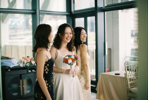Bride in white dress holds colorful bouquet, laughing with two bridesmaids in elegant venue.