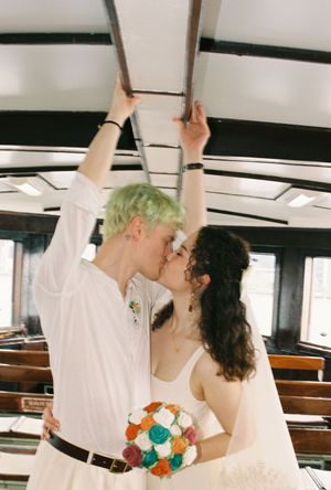 Couple kissing on a vintage trolley, bride holding colorful bouquet, groom with green hair in white shirt.