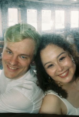 A smiling couple poses together in a bright restaurant, the man in a white shirt and woman with curly hair wearing a white top.