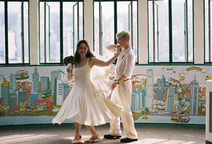 Couple dancing in white outfits before colorful city mural with large windows in background.