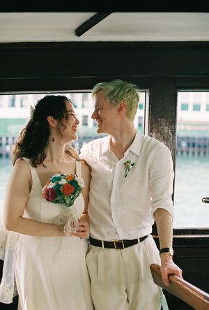 A couple in white wedding attire smiles at each other, the bride holding a colorful bouquet aboard a waterfront venue.